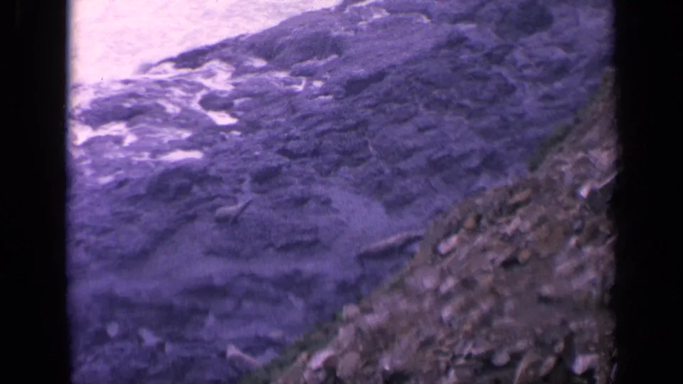 Vintage 8mm view of California coastal surf breaking over dark rocks, with loose stones along the shore.