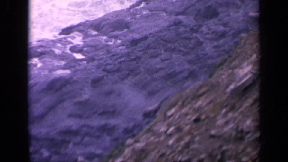 1963 coastal home-movie frame with waves and foam crossing dark rock formations beside a sloped rocky bank.