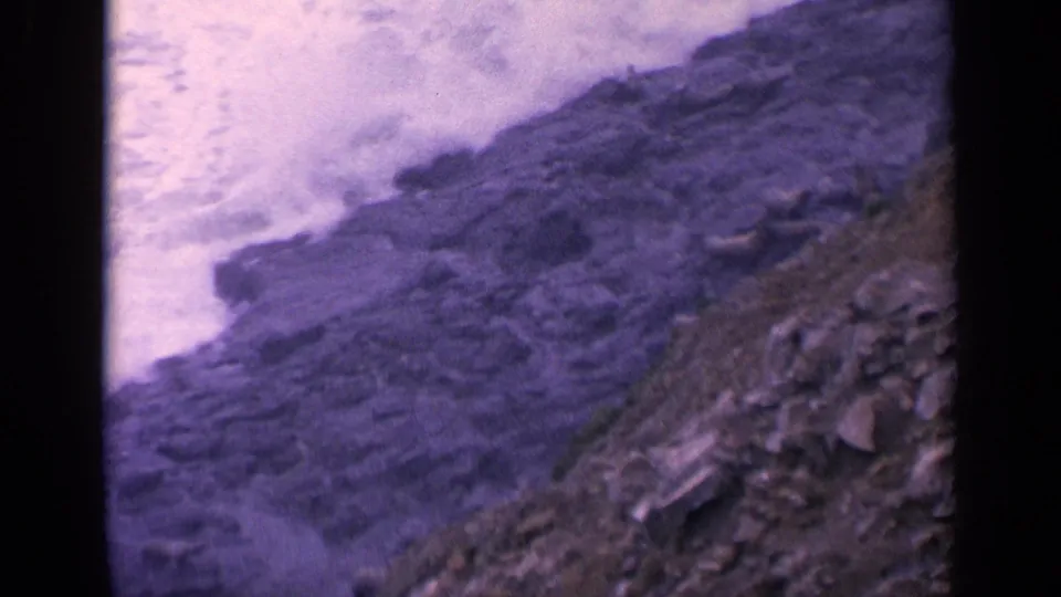 California 8mm frame from 1963 showing pale surf breaking against dark coastal rocks and a rocky shore edge.