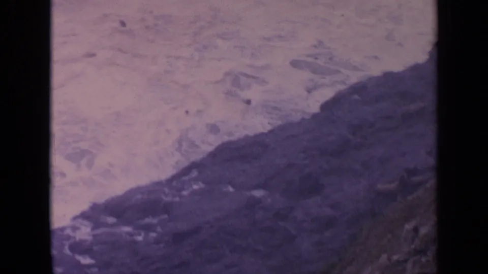 1963 California home-movie frame of surf and foam pushing against a dark rocky shoreline ledge.
