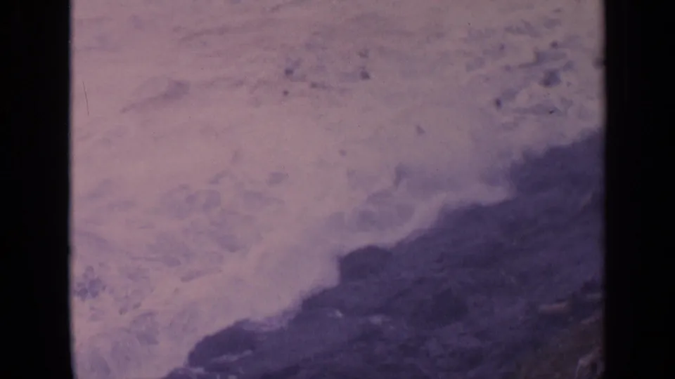 Soft 8mm frame of California surf spreading over rocks with a dark coastal edge in the foreground.