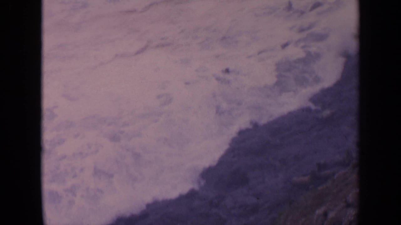 1963 California 8mm frame showing pale surf and churning water near dark coastal rocks.
