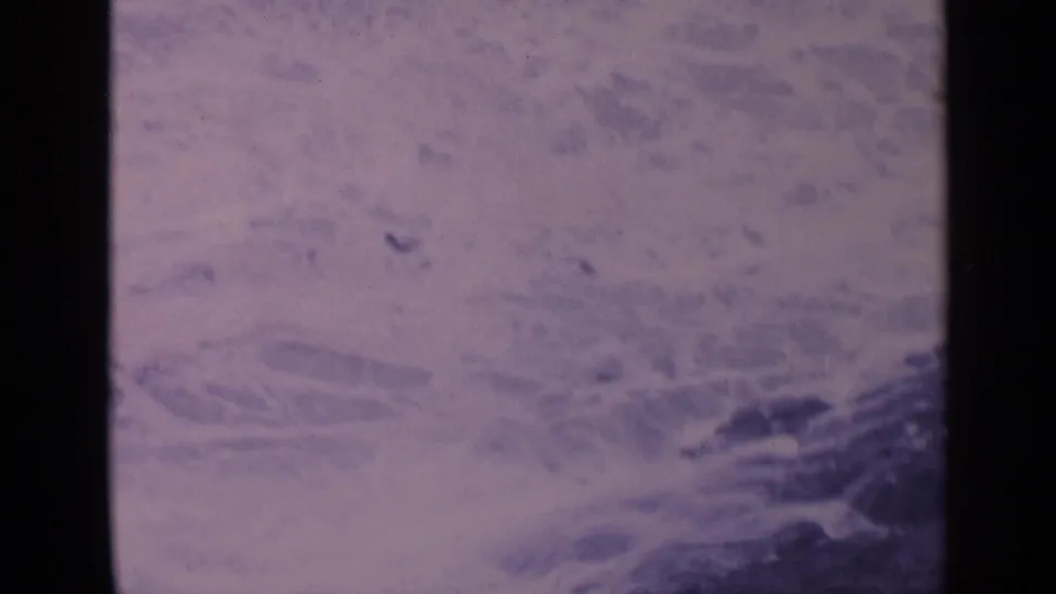1963 California 8mm footage shows foamy ocean water moving around dark rocks, framed softly with visible film grain and edge shadow.