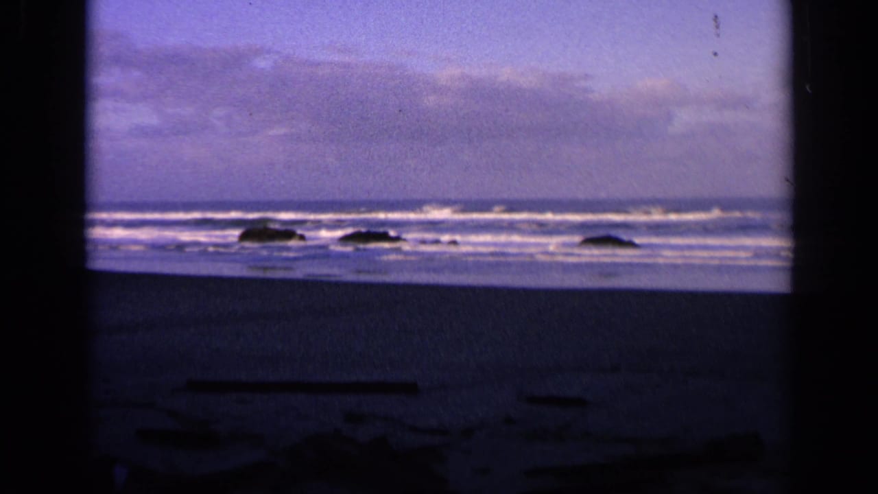 1963 California 8mm frame showing surf, white foam, dark rocks, and a dim beach foreground at dusk.