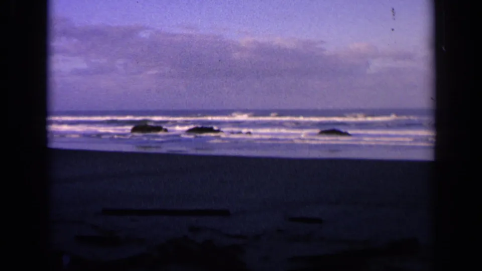 1963 California 8mm frame showing surf, white foam, dark rocks, and a dim beach foreground at dusk.