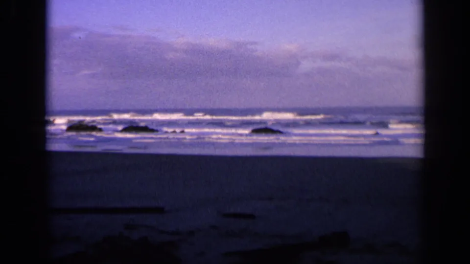 1963 California 8mm frame with ocean foam rolling toward scattered rocks and a shadowed beach foreground.