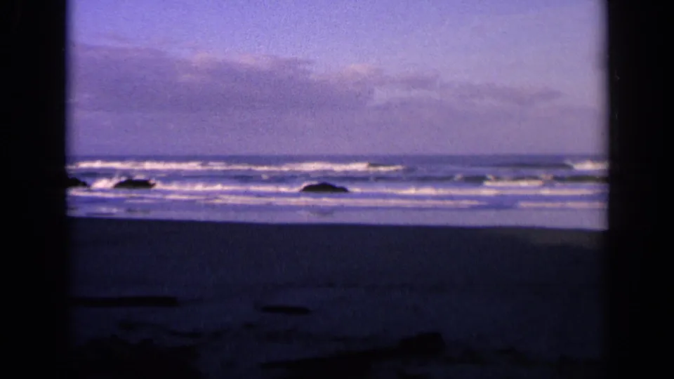 1963 California 8mm frame showing a wider band of surf, dark shoreline rocks, and soft twilight color.