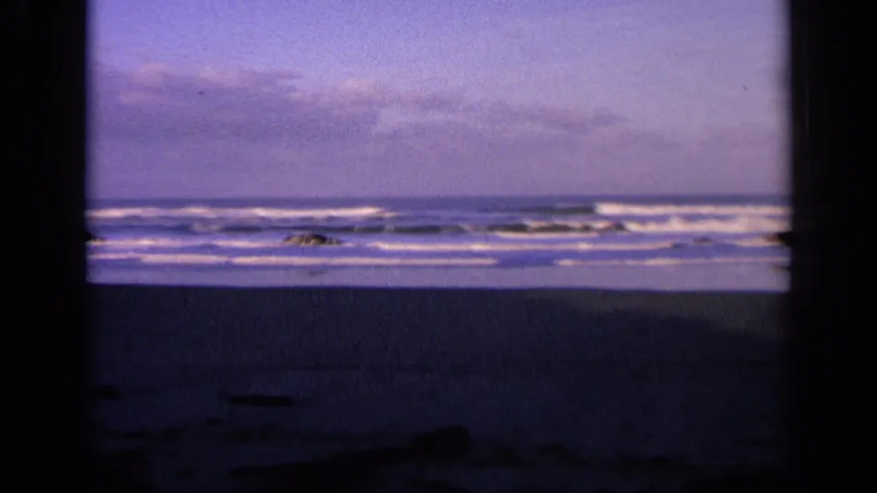1963 California 8mm frame with foamy ocean waves, low rocks, and a dark foreground at the end of the pan.