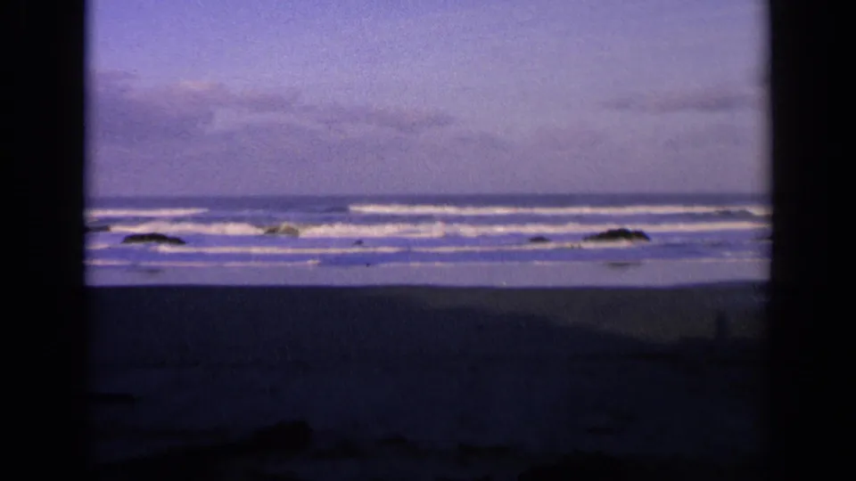1963 California coastal frame showing low waves, scattered rocks, wet beach, and a purple-toned sky.