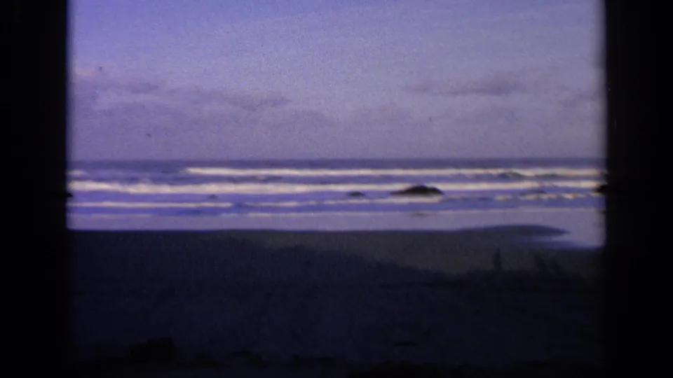 1963 California home-movie frame of a broad shoreline, white surf bands, rocks, and muted blue-purple sky.