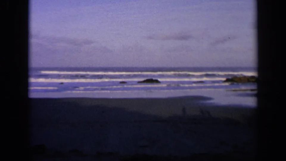 1963 California 8mm frame of waves breaking near rocks, with wet sand and dark edge framing in the foreground.