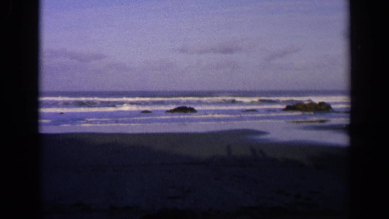 1963 California 8mm frame of a deserted beach at dusk, with surf bands, shoreline rocks, wet sand, and dark edge vignetting.
