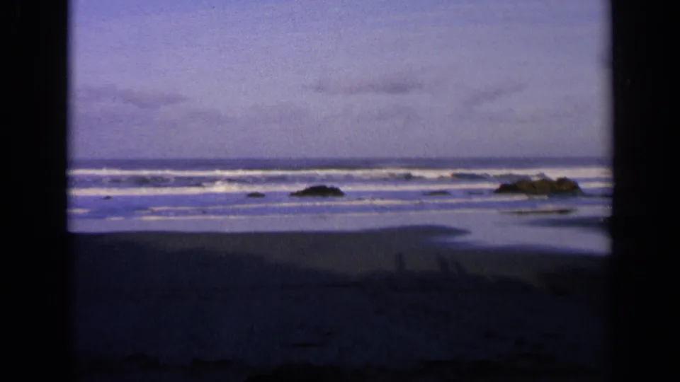 1963 California 8mm frame of a deserted beach at dusk, with surf bands, shoreline rocks, wet sand, and dark edge vignetting.