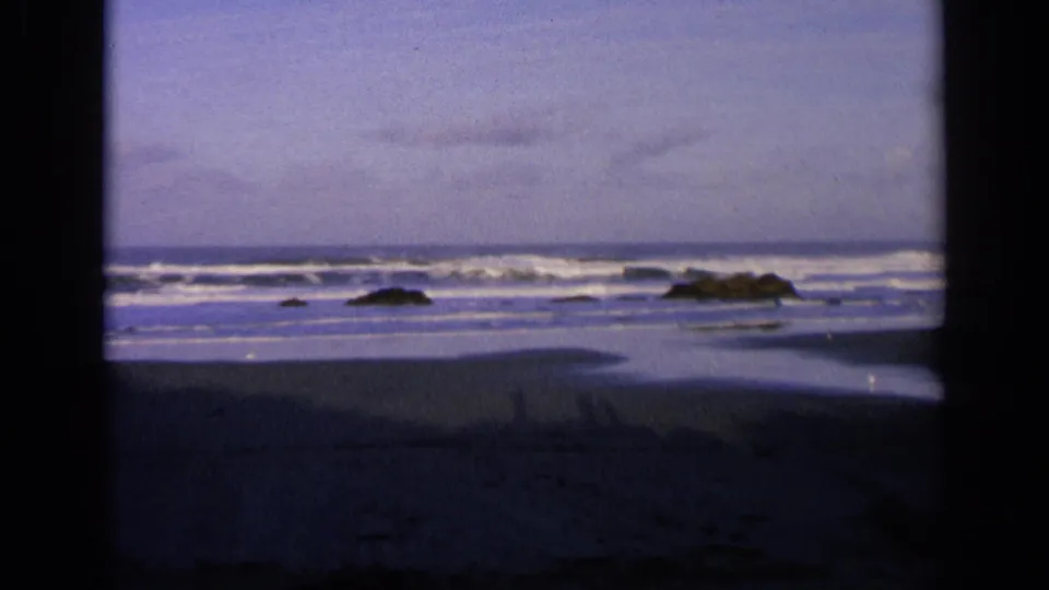 1963 California coastal home-movie frame showing dusk surf, low rocks, wet beach sand, and a blue-purple sky.