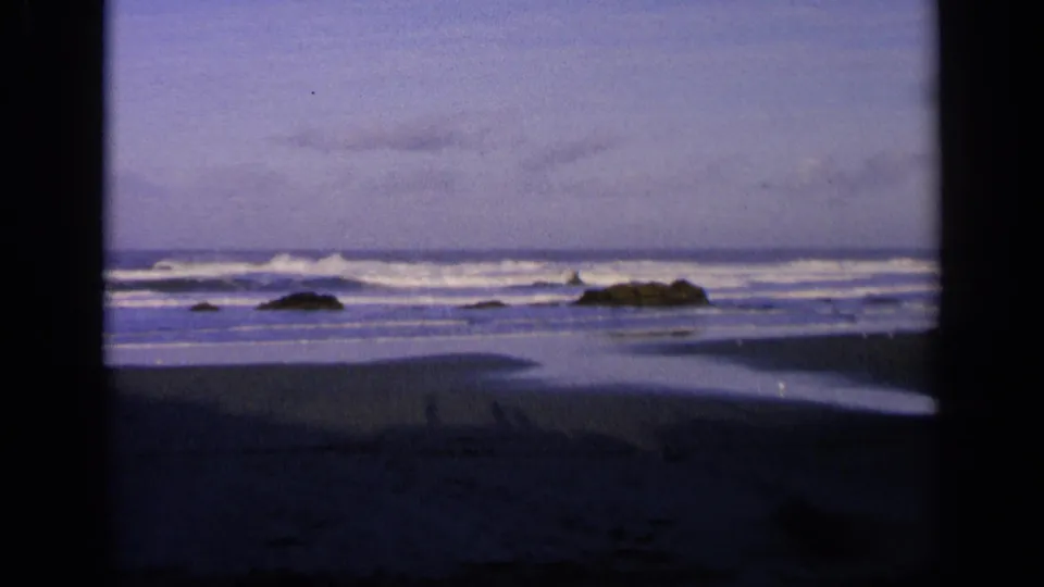 1963 California 8mm view of waves breaking at dusk, with scattered dark rocks, wet sand, and soft grainy color.