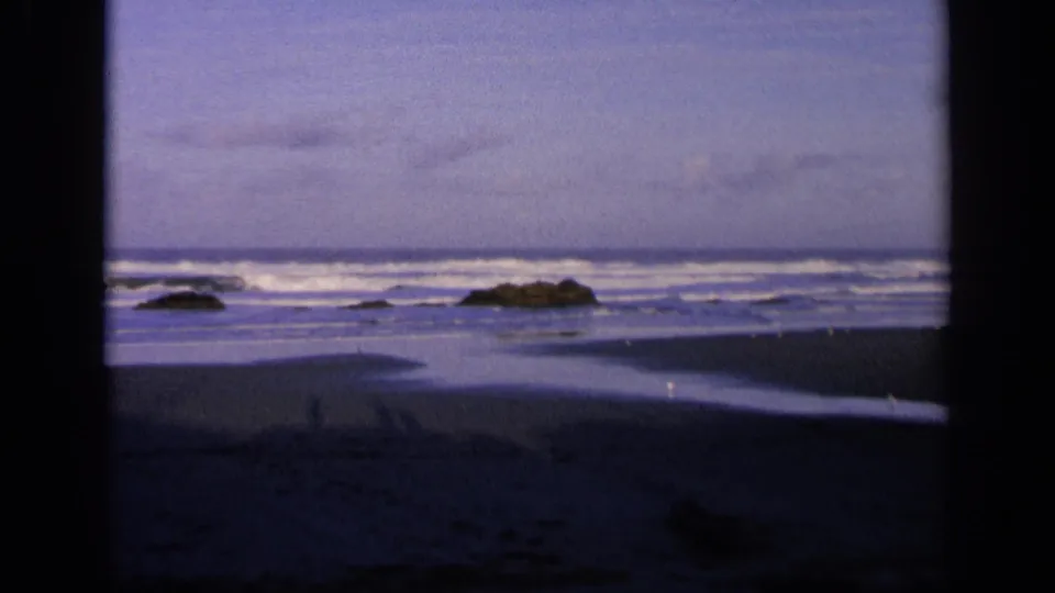 1963 California beach frame with distant surf, low rocks in the water, wet sand patterns, and pronounced 8mm vignetting.