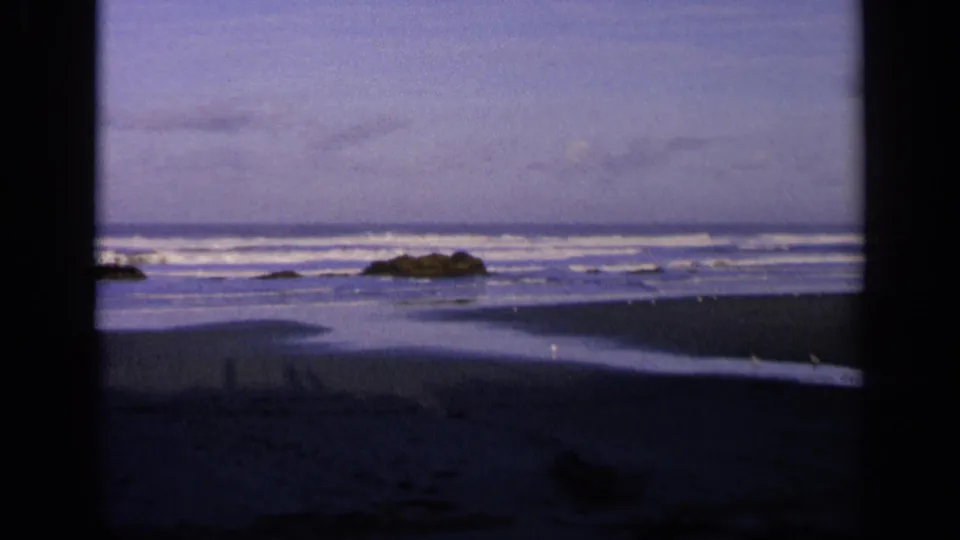 1963 8mm home-movie view of a California shoreline through a dark frame, with surf, wet sand, birds, and low rocks under blue sky.