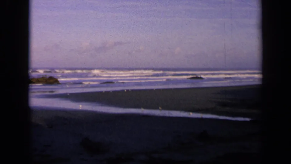 Final 1963 California coast still shows a quiet beach through dark side framing, with low surf, wet sand, birds, rocks, and a blue sky.