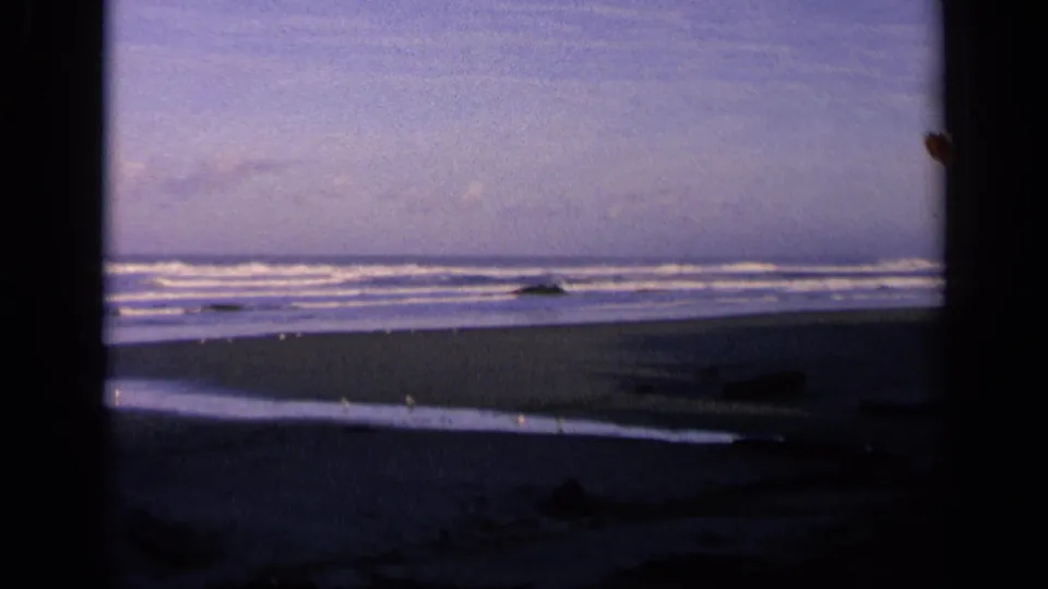 1963 California coastal home-movie frame with pale surf lines, empty sand, offshore rocks, and a heavy dark border at the left and center.