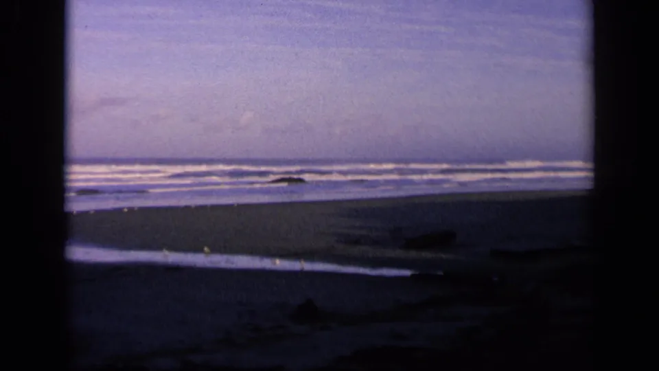 1963 California 8mm still of an empty shoreline, with gentle waves, dark foreground rocks or driftwood, and a shaded border around the view.