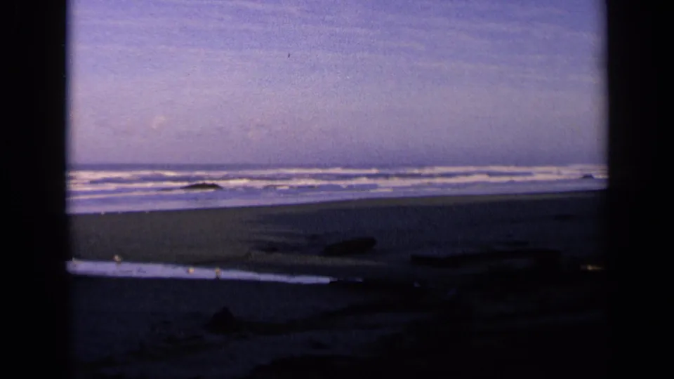 1963 California beach frame with subdued color, low surf, scattered shoreline debris, and a dark foreground edge suggesting a window or doorway view.