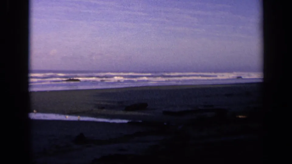 1963 California 8mm frame showing surf and a dim shoreline viewed through dark vertical framing, with driftwood on the beach.