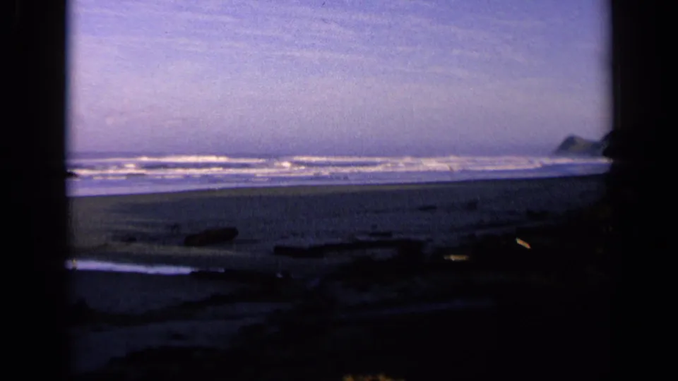 1963 California 8mm still of waves and a distant rocky point, with dark foreground framing and driftwood on the sand.