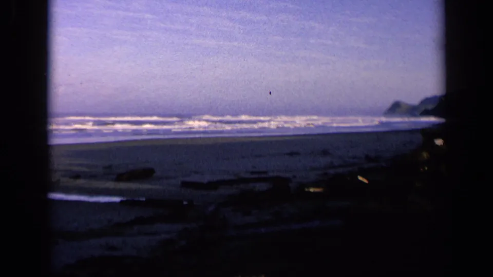 Vintage 8mm coastal frame showing California surf and dark beach foreground, with distant rocks under a blue-purple sky.