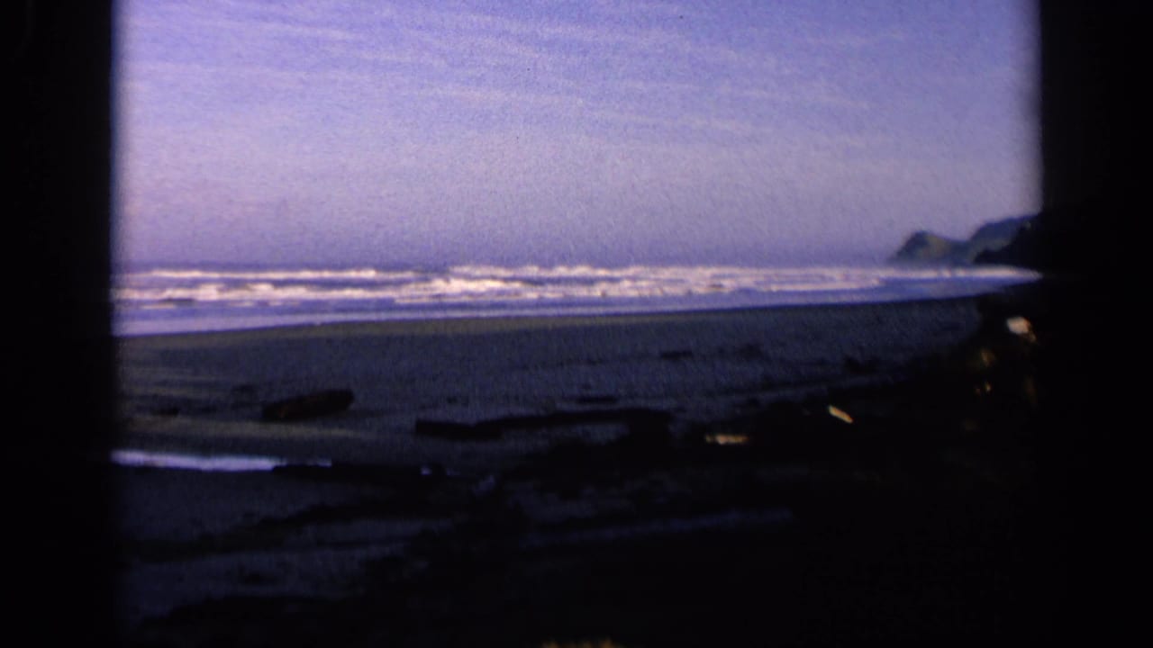 1963 California 8mm frame showing a wide shoreline with waves, dark beach debris, wet sand, and distant coastal rocks.