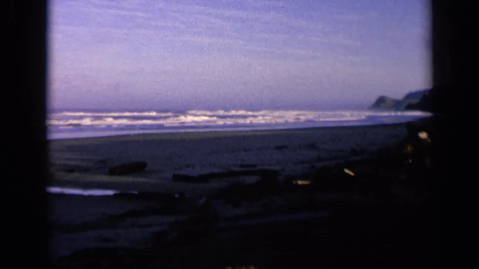 1963 California 8mm frame showing a wide shoreline with waves, dark beach debris, wet sand, and distant coastal rocks.