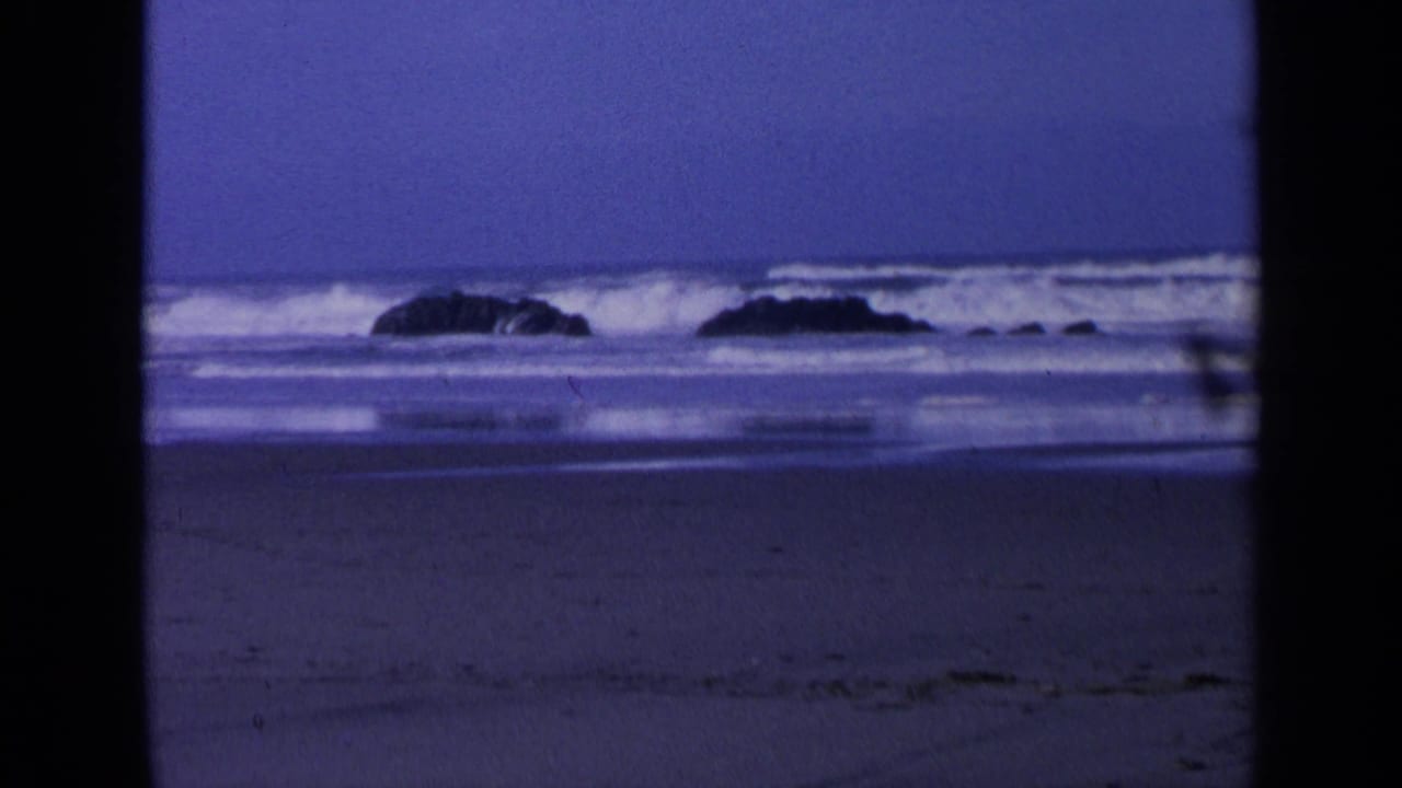 Dark boulders offshore with waves breaking behind them and wet sand in the foreground.