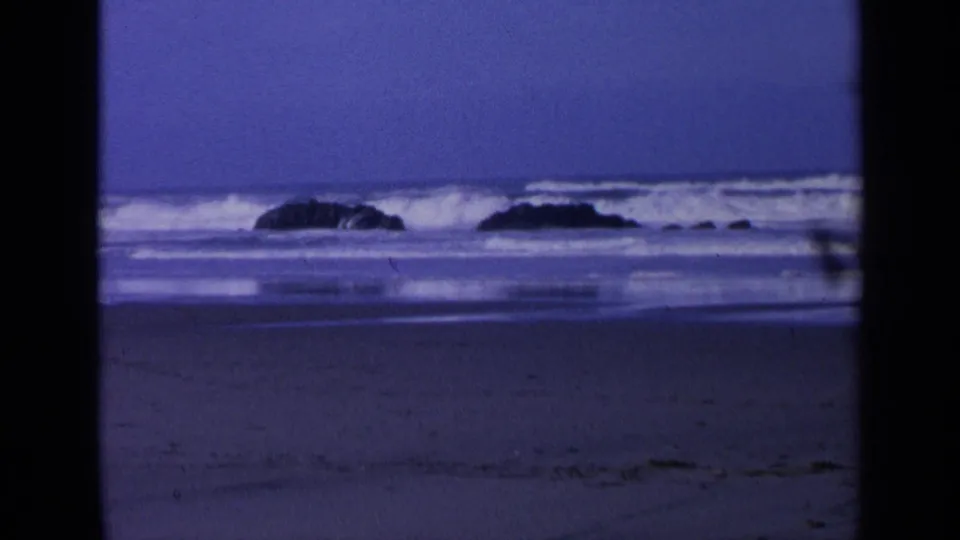 1963 California 8mm beach view with dark offshore boulders, rolling surf, wet sand, and a cool purple-blue cast.