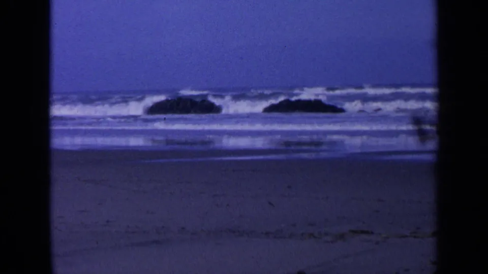 A 1963 home-movie frame of a California beach with offshore rocks, white surf, wet sand, and visible film grain.
