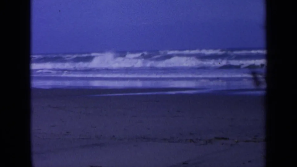 Final 1963 California beach frame with a breaking wave, distant rocks, wet sand, and aged 8mm color shift.