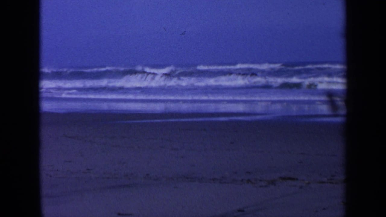 Blue-toned surf breaks near dark rocks on a sandy beach.