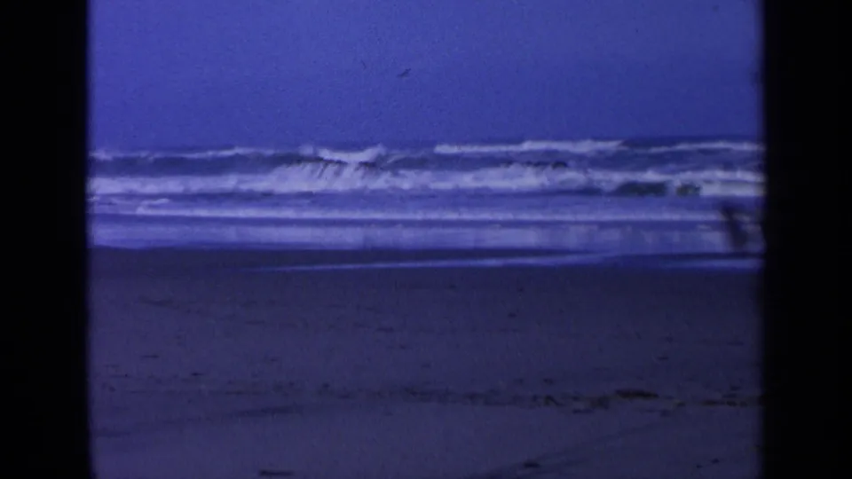 1963 California 8mm frame showing white ocean waves breaking near dark rocks under a deep blue sky.