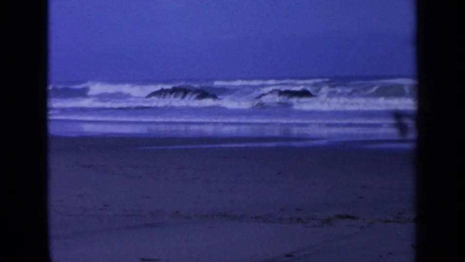 Blue-cast 8mm view of California surf rolling around two dark rocks near the sandy shore.