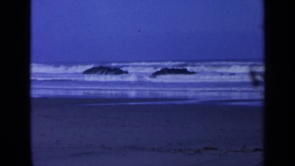 1963 California home-movie frame with low white surf crossing the shore and dark rocks in the water.