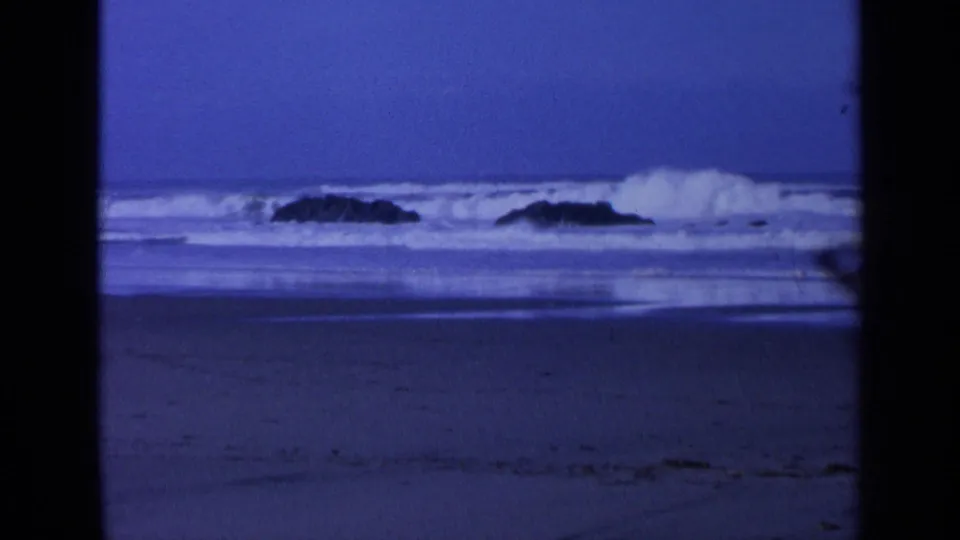 Blue-toned 8mm frame of a breaking California wave throwing white foam beside dark rocks near shore.