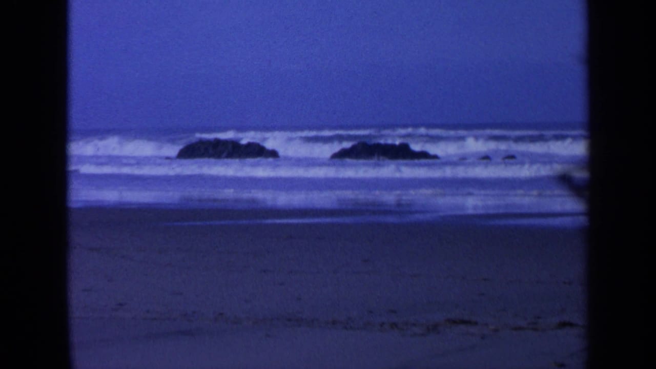 Dusk ocean surf breaking around dark rocks beyond a wet beach.