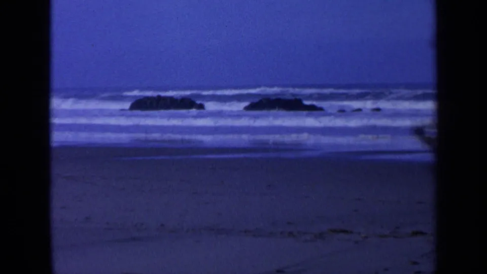 Blue 8mm view of California surf, offshore rocks, and a dark beach under a dusk sky.