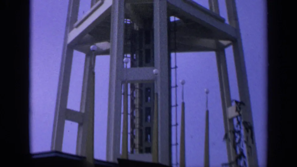 1963 California 8mm frame showing the tower's angled supports, central vertical core, and slender spires against a purple sky.