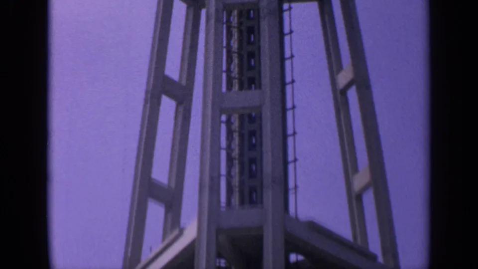 1963 California 8mm frame showing the lower-to-middle portion of a tall steel tower with ladder and bracing against a purple sky.