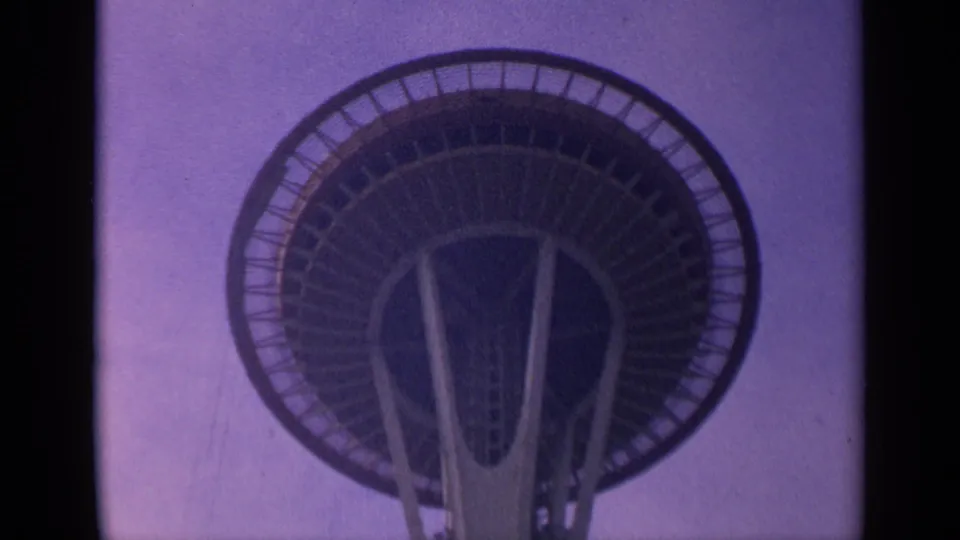 Low-angle 1963 8mm view of the Space Needle's saucer-shaped deck and central structure, framed by dark scan borders.