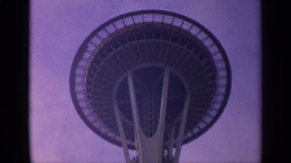 1963 home-movie frame of the Space Needle viewed upward, with the circular deck, railings, and angled supports prominent.