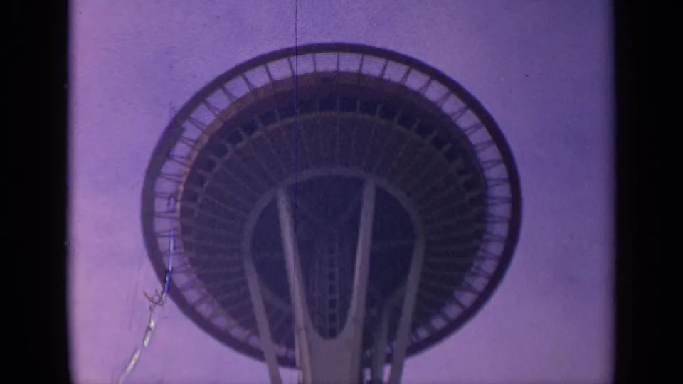 Final 1963 8mm frame of Seattle's Space Needle from below, with the observation deck, support legs, and a thin foreground pole or wire visible.