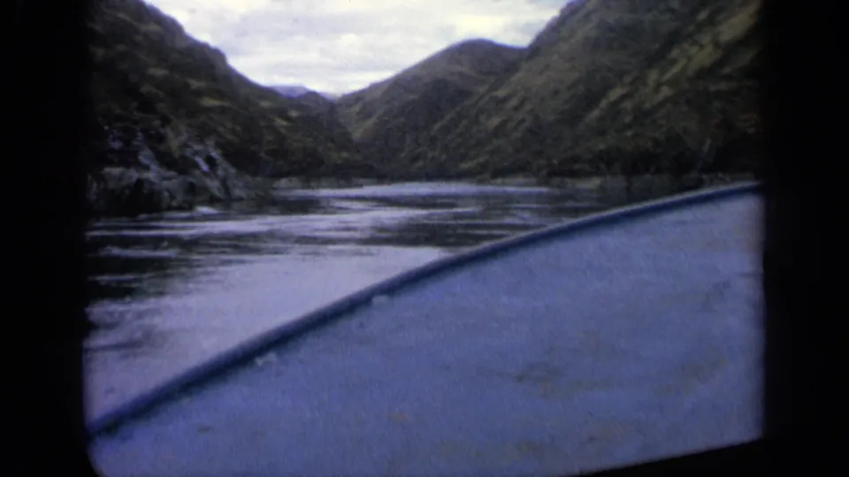 1963 home-movie frame from a boat traveling along an Idaho river bordered by steep, shadowed hills.
