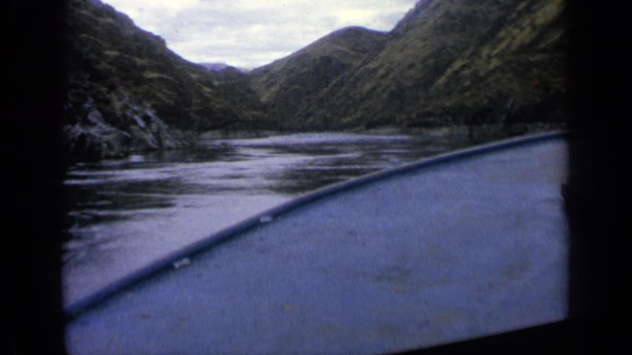 1963 Idaho 8mm frame showing a boat-level view across calm river water toward steep green mountain slopes and an overcast sky.