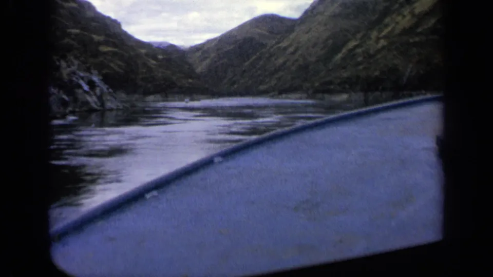 1963 Idaho home-movie frame from a boat, looking across reflective river water toward a narrow mountain valley.
