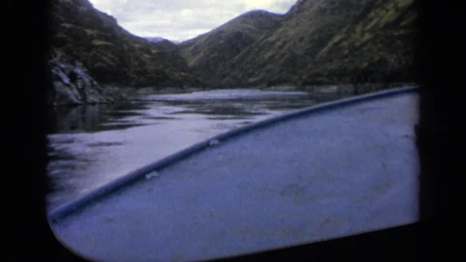 1963 Idaho 8mm still with the boat edge crossing the foreground, calm water, a rocky bank, and steep wooded slopes beyond.